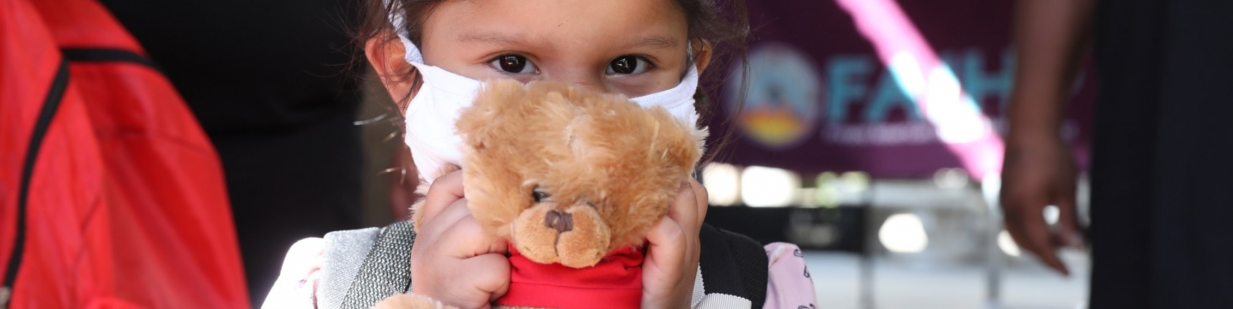 bimba con capelli castano scuro, bianca di carnagione, indossa una mascherina per proteggersi dal covid-19 e tiene tra le mani di fronte al viso un peluche a forma di orsacchiotto