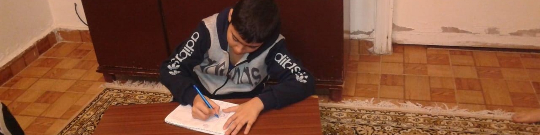 Bambino libanese di circa 12 anni è seduto a terra su un tappeto e scrive su un figlio bianco appoggiato a una tavolo di legno.