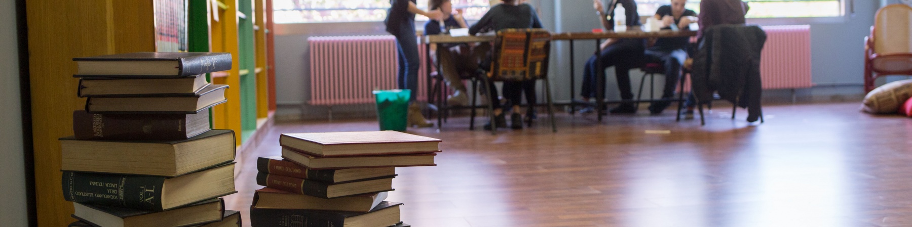 biblioteca, in primo piano dei libri accatastati e sullo sfondo delle persone intorno a un tavolo