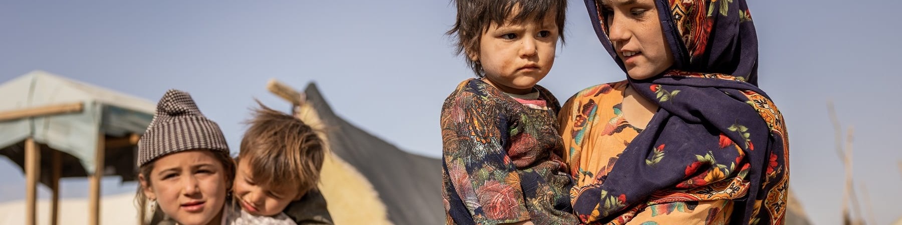 Quattro bambini afgani: una bambina tiene in braccio un bambino più piccolo, un altro bambino tiene sulle spalle un bimbo piccolo. Sullo sfondo le tende del loro villaggio