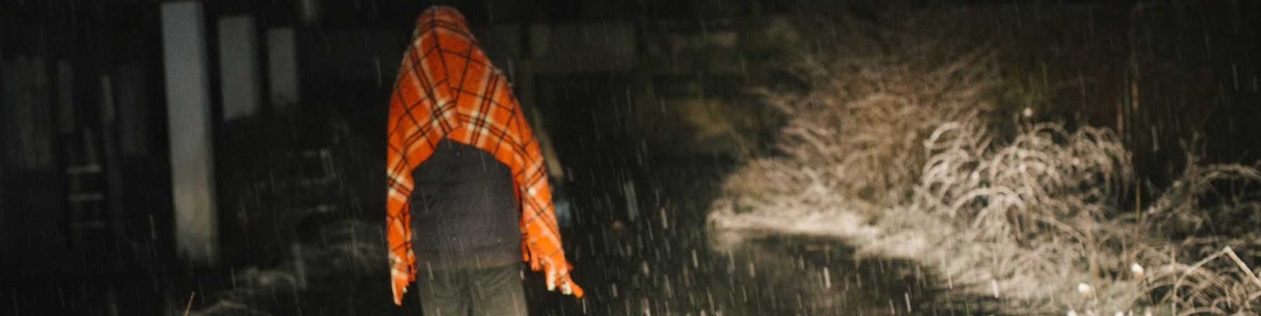 Foto notturna in un viale oscuro mentre sta nevicando. Si vede di spalle un ragazzo che cammina lungo il viale e sotto la neve con pantaloni scuri e una coperta arancione posta sopra le spalle e la testa.