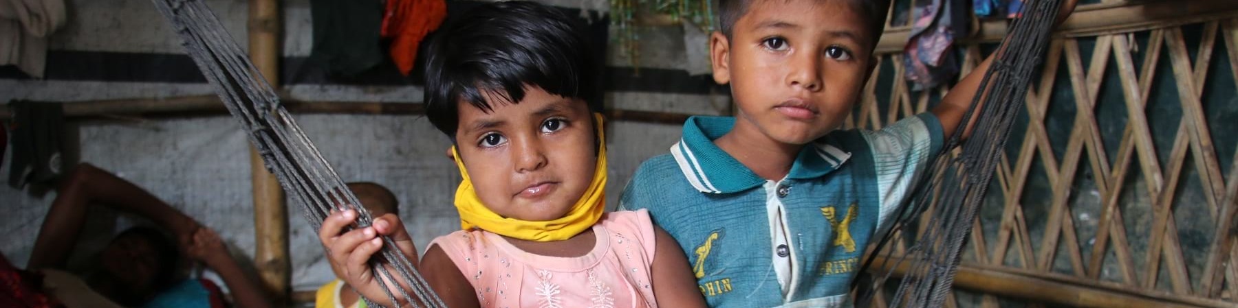 Due bambini Rohingya sono seudti a terra vicini. Il maschio è piu grande e indossa jeans lunghi e una maglietta a maniche corte verde, la bambina ha una canottierina bianca e rossa e una gonna e tiene in mano un peluche giallo