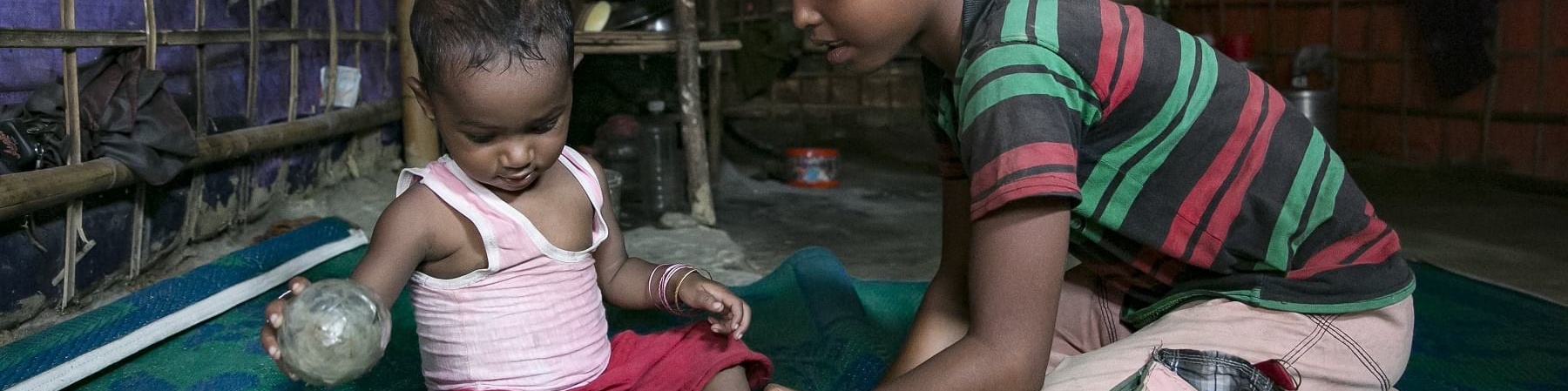 Un bambino di circa sei anni gioca seduto a terra su un tappeto verde con un bambino di circa un anno. Entrambi sono del Myanmar.