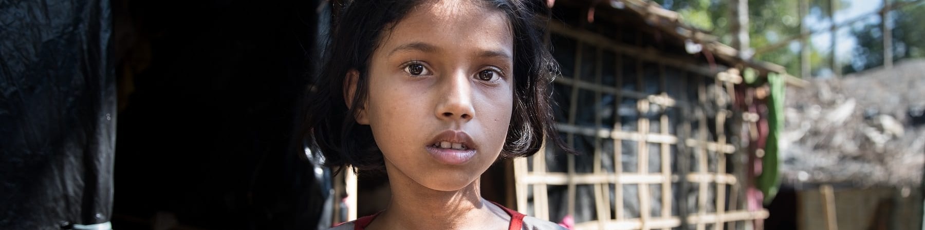 Primo piano di una bambina bengalese mora con capelli corti, la bocca leggermente aperta. Indossa una maglietta grigia con righine rosse.