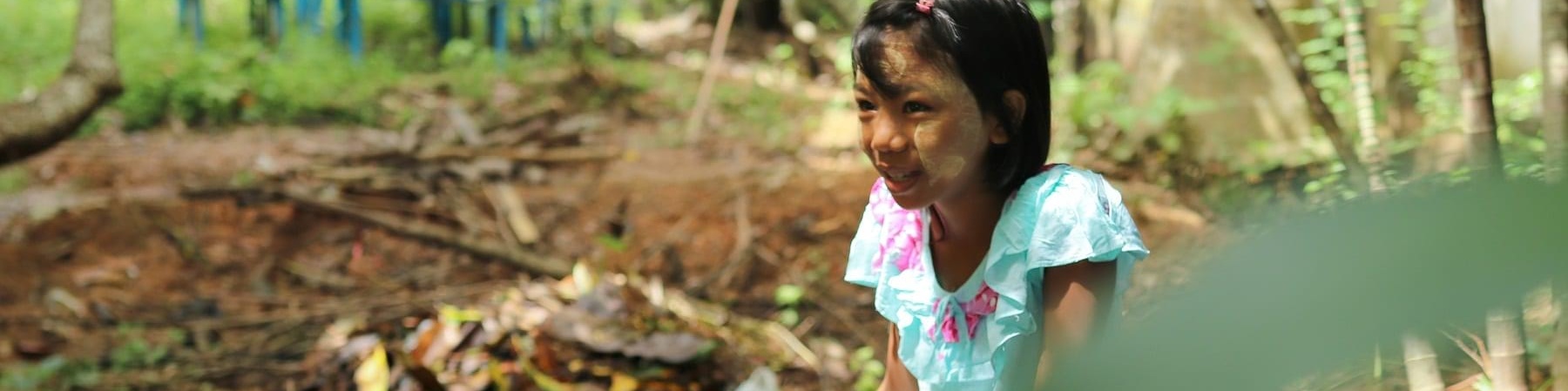 bambina del myanmar con vestito celeste a fiori rosa è seduta su una sedia in mezzo a un terreno circondata da piante