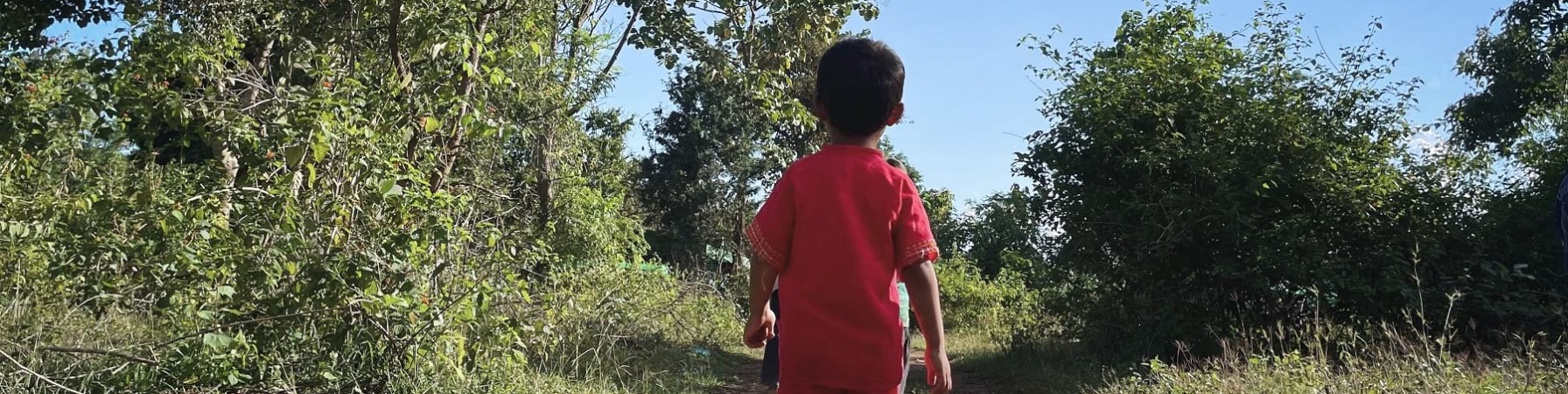 un bambino di circa 5 anni di spalle vestito con maglietta a maniche corte e pantaloncini rossi cammina con infradito in una strada sterrata di un campo sfollati in myanmar