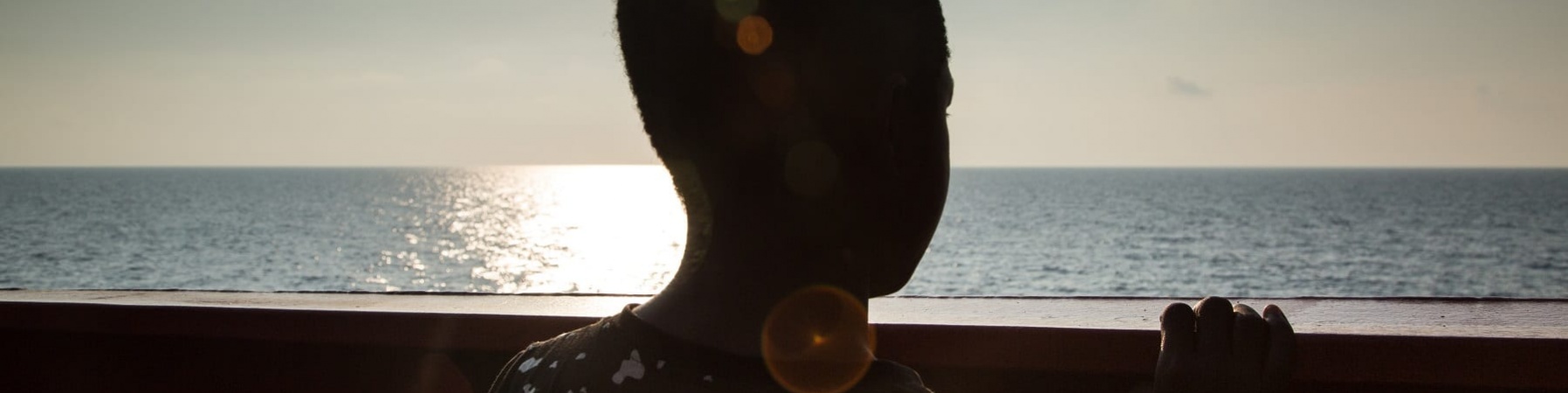 Un bambino contro sole di spalle è affacciato alla balaustra di una nave e guarda il mare
