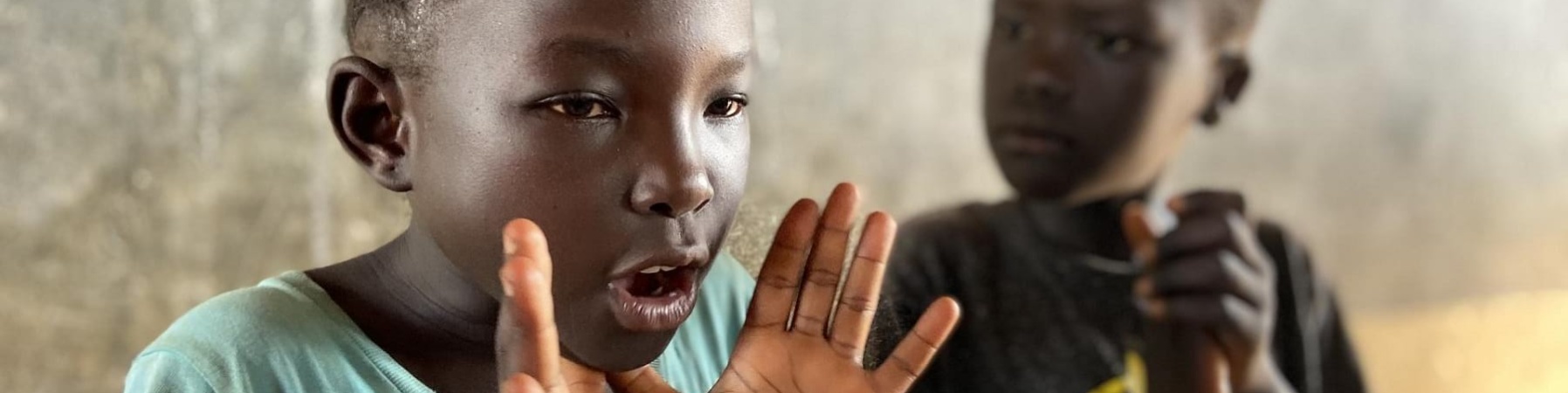 Un bambino nero sullo sfondo batte le mani mentre un bambino nero in primo piano fa un gesto con la mano poggiando i pollici sotto il mento con la mano aperta