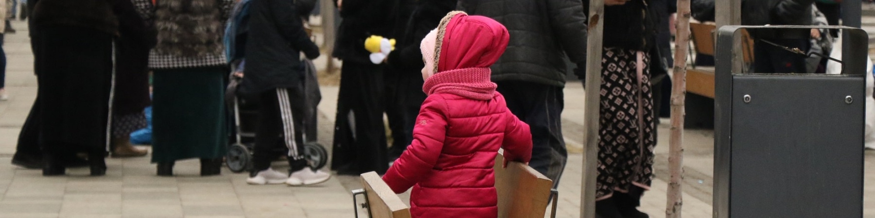 bambina con giacca rosa sola seduta su una panchina, sullo sfondo una folla di persone con trolley e zaini che fugge