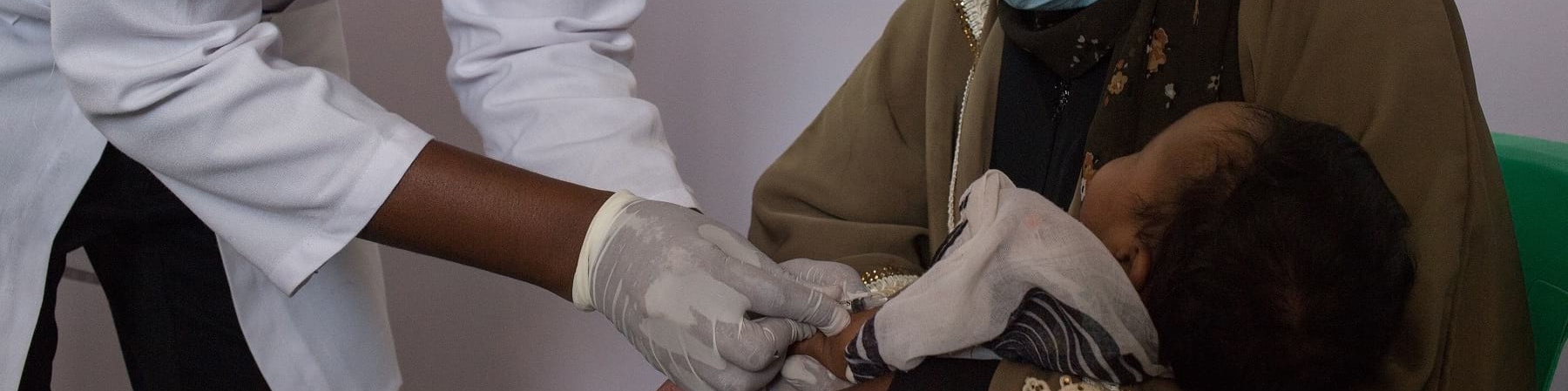 Un operatore save the children con indosso camice e mascherina vaccina un neonato in braccio a sua madre anche lei con maschrina