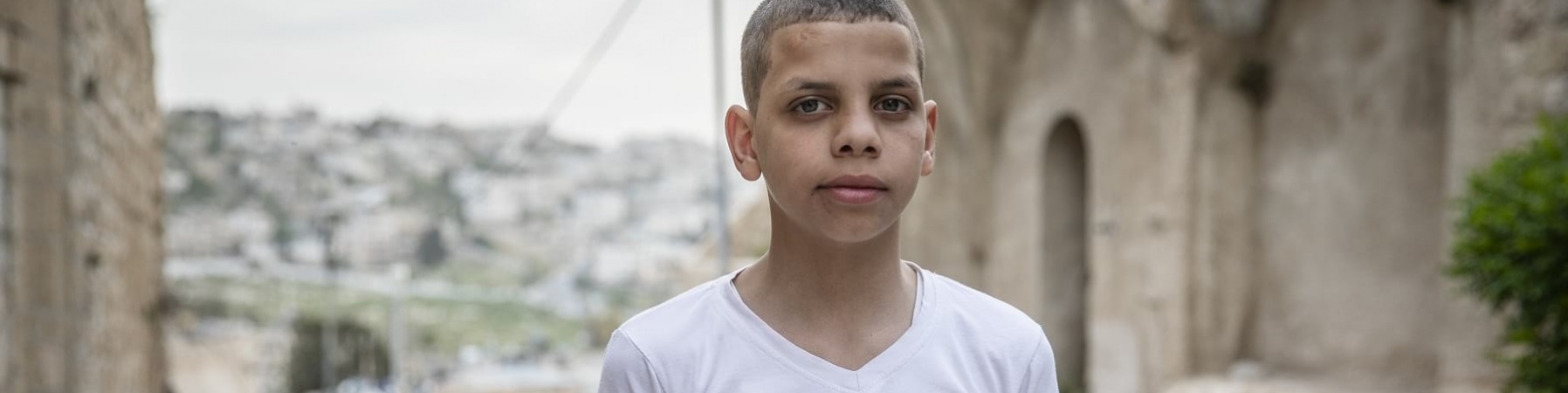 bambino dei territori palestinesi occupati ripreso a mezzo busto con maglietta bianca e sfondo paesaggio sfocato