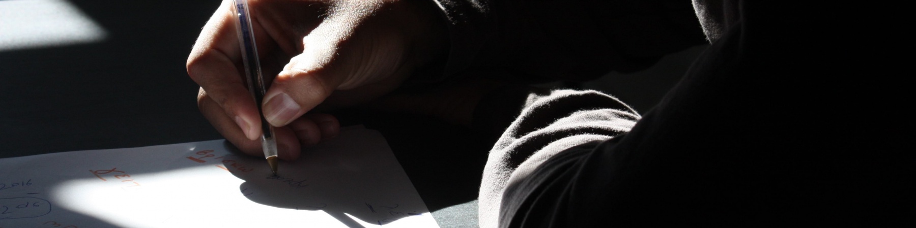 Braccia di un bambino poggiate su un tavolo, una mano scrive con una penna su un foglio bianco. Tutta la foto è in penombra