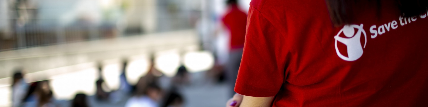 Operatrice save the children con la maglietta ripresa di spalle fino a sotto la vita, guarda bambini seduti a terra ma sfocati nella foto