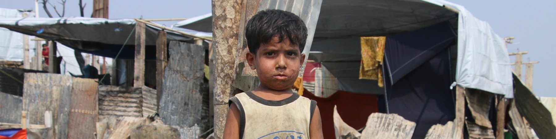 bimbo Rohingya in un campo profughi, vestito con canottiera e pantaloncini sporchi di terra, sullo sfondo un rifugio di lamiere fatiscente
