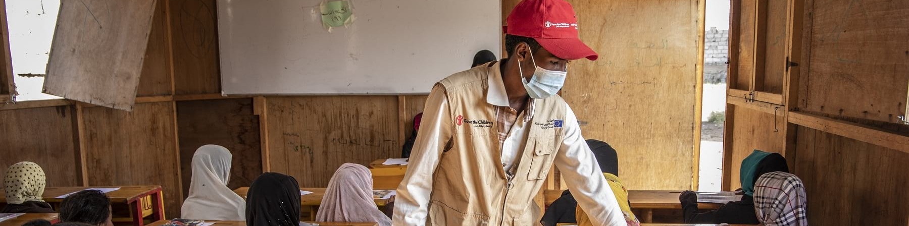 Un operatore save the children in piedi tra i banchi dei bambini in un aula scolastica di un campo rifugiati in Yemen