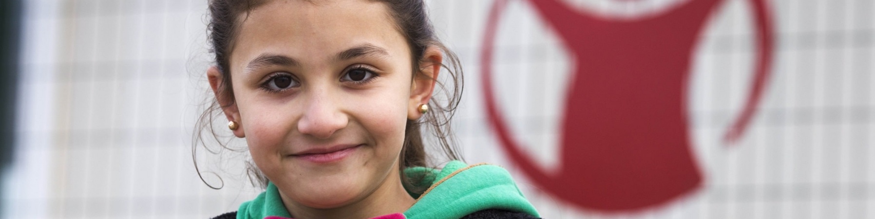 bambina sorridente con maglioncino verde e giacca, sullo sfondo il logo di save the children