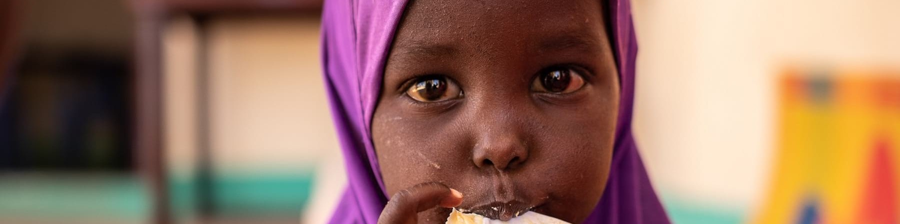 Bambina somala di pochi anni con chador viola in testa mangia una con le mani da una bustina di plumpynat