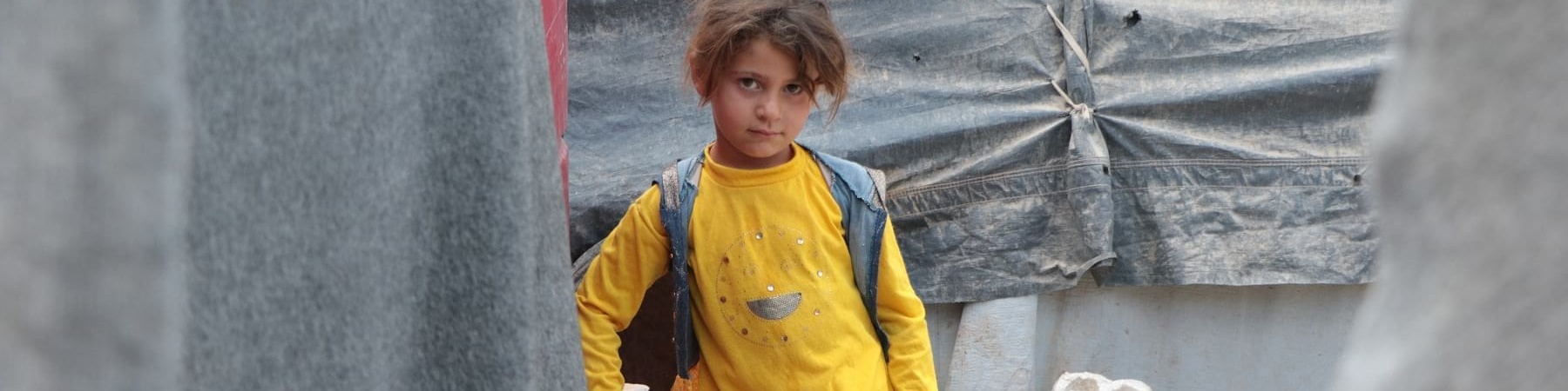 Una bambina vestita di giallo cammina in un campo profughi di cui si vedono alcune parti delle tende e dei rifiuti ingombranti dietro di lei.