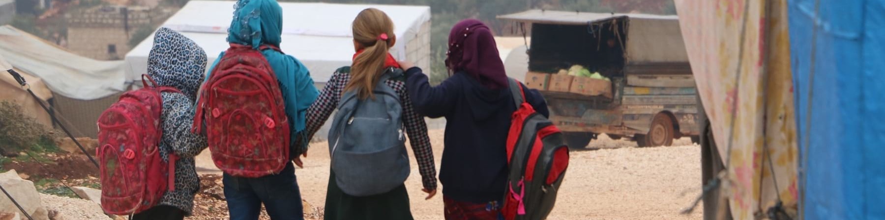 quattro bambini siriani camminano di spalle con gli zaini sulle spalle in un campo per sfollati
