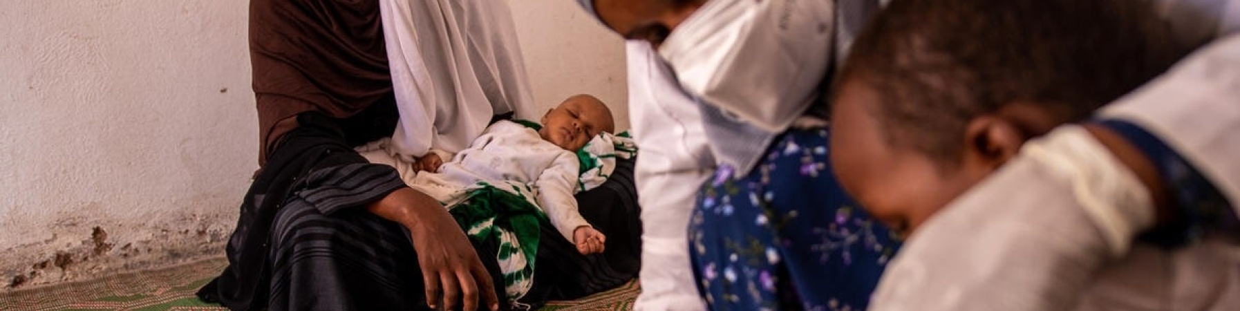 bambino somalo in primo piano mentre gli viene misurato dall operatrice la malnutrizione e sullo sfondo donna con bambino piccolo sulle gambe 