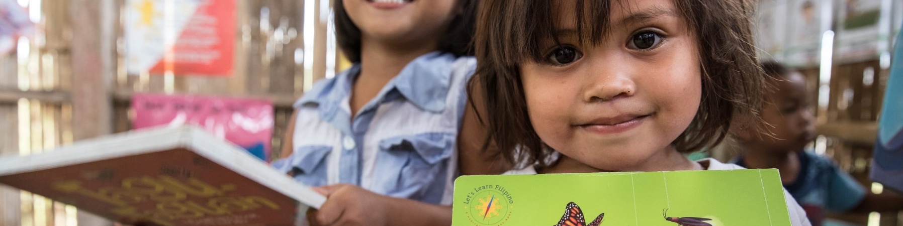 due bambine dentro un aula di scuola tengono in mano un libro a testa. Una di loro guarda in camera sorridente e laltra, sullo sfondo, ride.