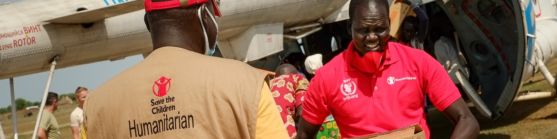 Operatori save the children trasportano in mano scatole di aiuti umanitari appena arrivati da un aereo. Un operatore è di spalle e l altro è di fronte.