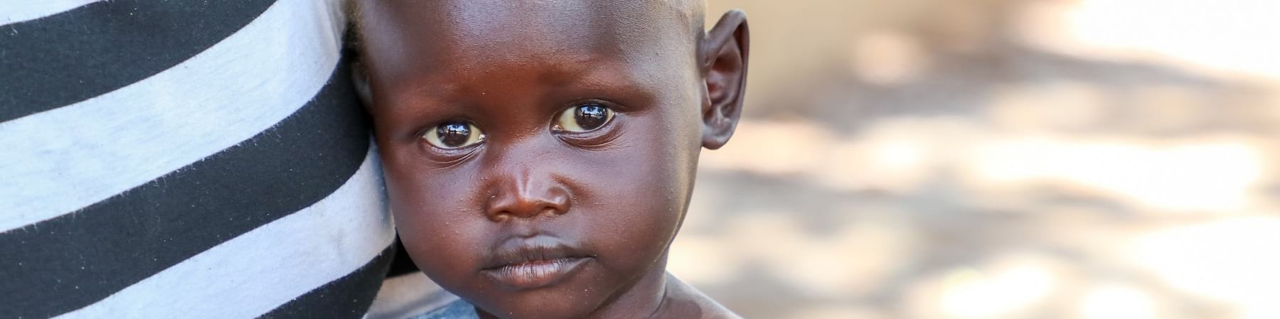 primo piano di un bambino sud sudanese con maglietta grigia è vicino alla pancia di un adulto con maglietta a righe bianche e nere