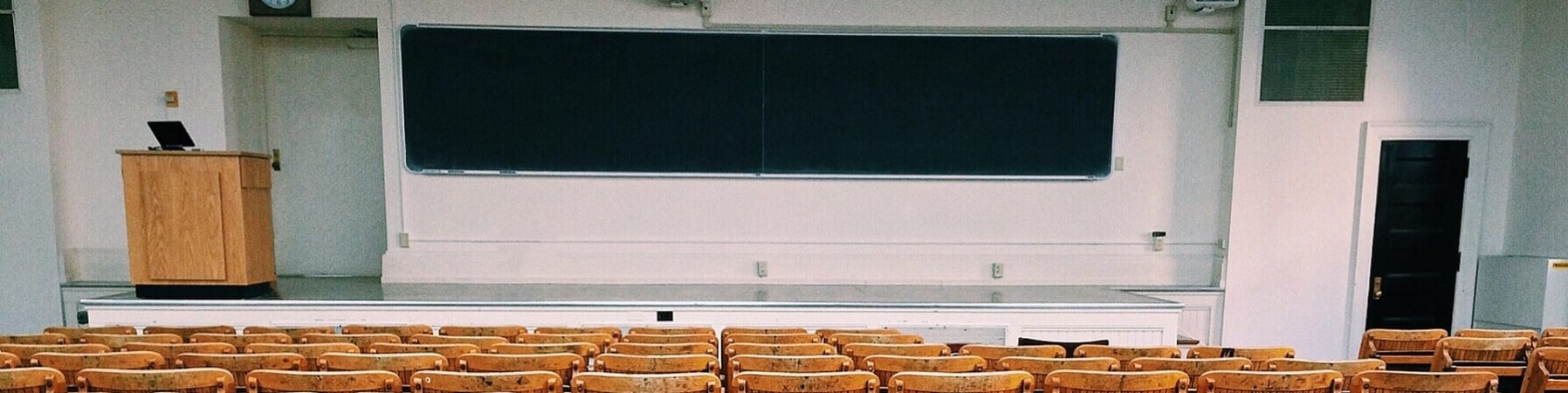 Aula universitaria vuota con sedie di legno pieghevoli, lavagna e leggio su un rialzo.