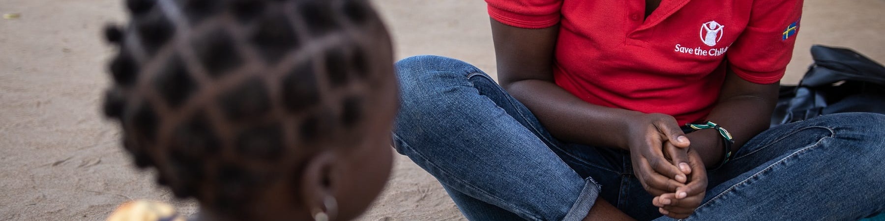 un operatrice save the children in Mozambico è seduta a terra a gambe incrociate e guarda una bambina di spalle seduta di fronte a lei.