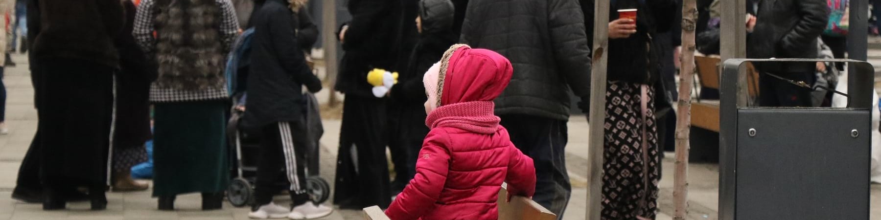 Una bambina con cappotto rosa di spalle è in ginocchio su una panchina. Davanti a lei una folla di persone.