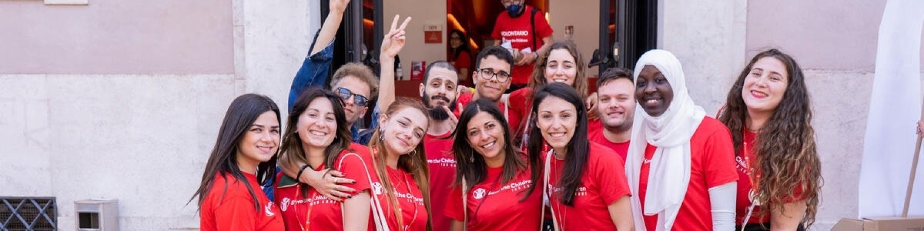 ragazzi e ragazze volontari di save the children che posano di fronte la sede sorridenti e con la maglietta rossa di save the children