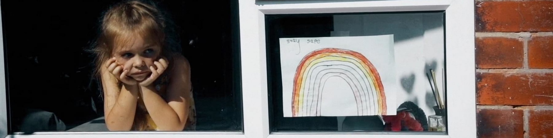 una bambina affacciata alla finestra guarda in basso pensosa. Sulla destra è appeso alla finestra il disegno di un arcobaleno