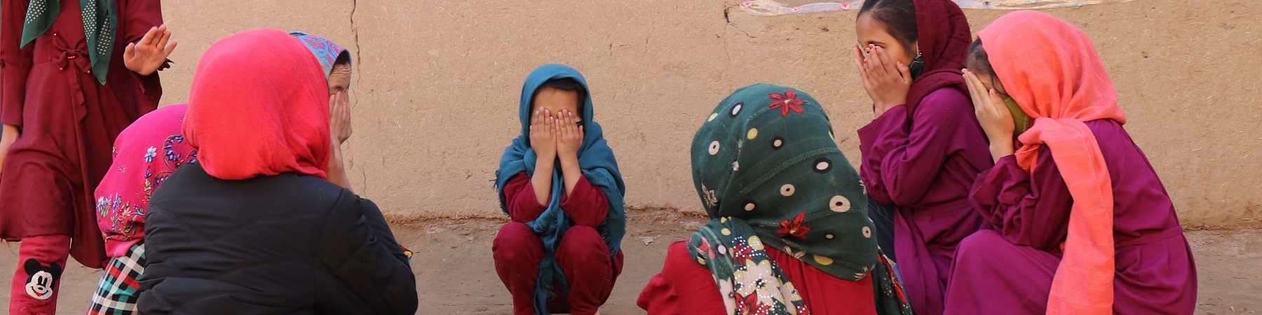 bambine vestite in viole e rosso sono sedute in cerchio e si coprono il volto con le mani.