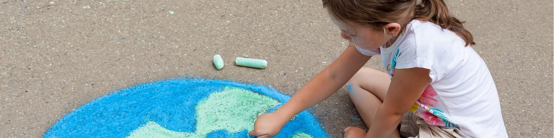 Una bambina bionda con la coda è seduta a terra sulla strada e disegna con i gessi un mappamondo. Lei è vestita con una maglietta a maniche corte bianca e dei pantaloncini marroni.