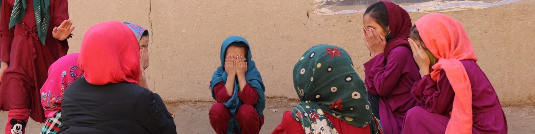 bambine vestite in viole e rosso sono sedute in cerchio e si coprono il volto con le mani.