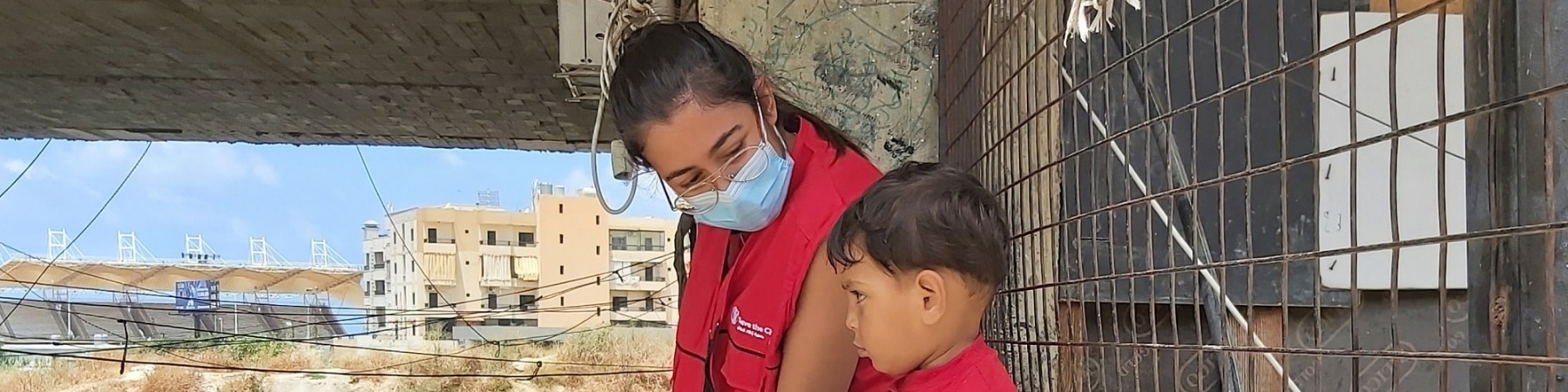 operatrice save the children con mascherina seduta su un muretto a fianco di un bimbo. Sullo sfondo varie presone che camminano. 
