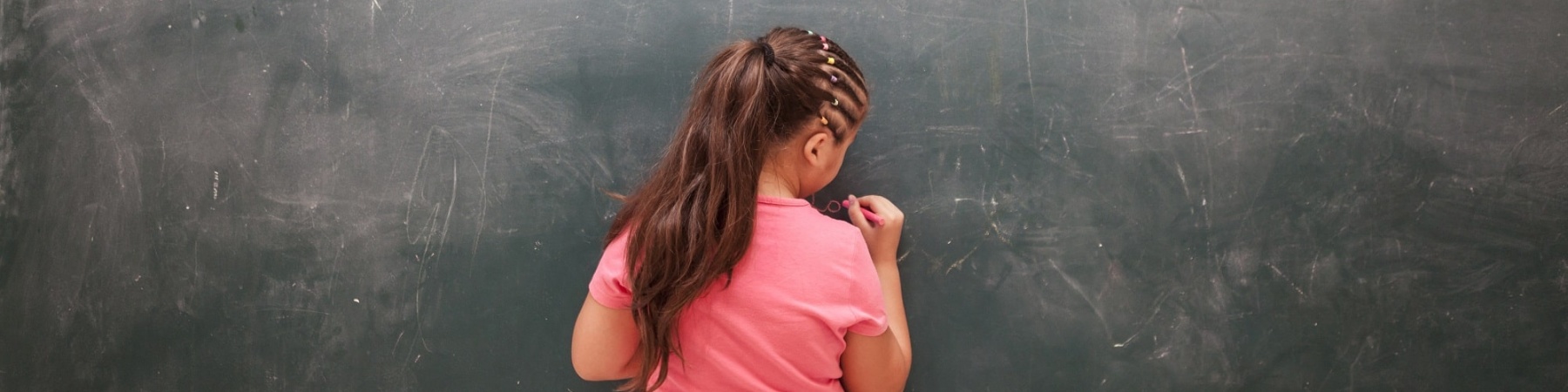 bambina di spalle davanti a una lavagna nera scrive con il gessetto