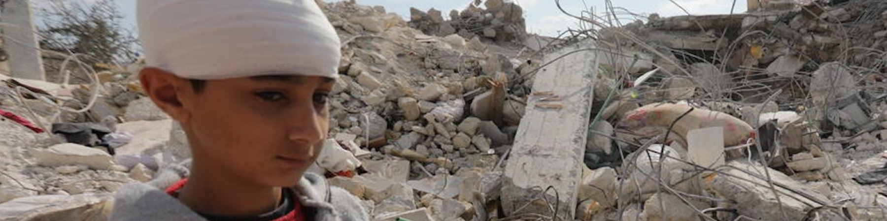 Un bambino con la testa fasciata guarda i resti distrutti della sua casa.