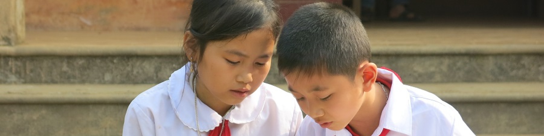Bambini con camicia bianca e cravattina rossa seduti su un muretto leggono un libro interessati