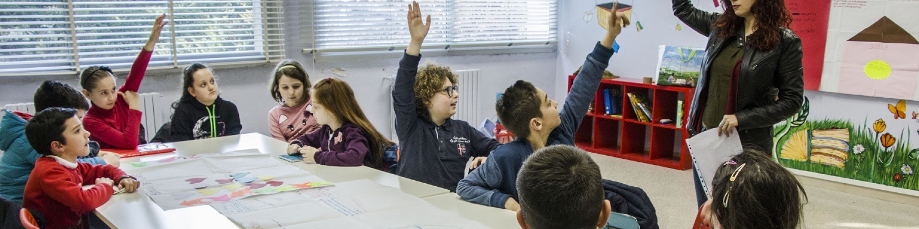una decina di bambini seduti a un banco di scuola alzano la mano alla domanda della maestra