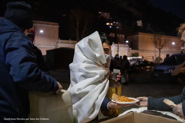 ragazzo migrante con una coperta addosso mentre riceve del cibo da un volontario