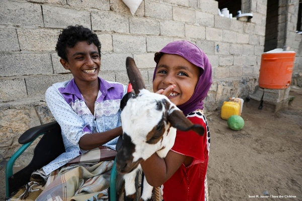 un bambino su una sedia a rotelle, davanti una bambina con una capretta in braccio, entrambi sorridenti fuori dalla loro casa, siamo in Yemen
