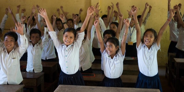 bambine e bambini tra i banchi di scuola con le mani in alto sorridenti 