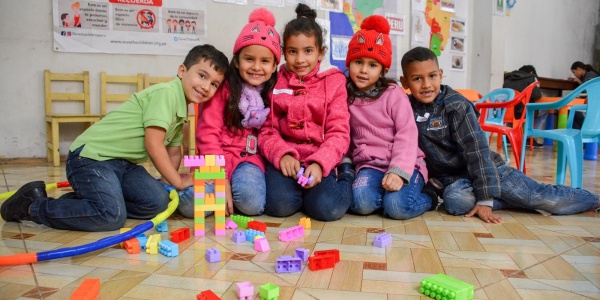 5 bambini seduti per terra a giocare in un asilo. Indossano maglioncini colorati