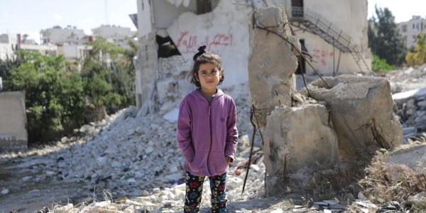 Bambina siriana in piedi tra le macerie. Indossa pantaloni neri e una felpa viola e tiene le mani nelle tasche
