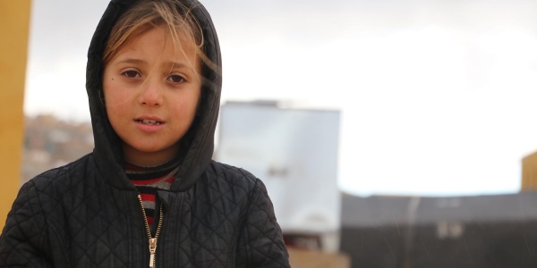 Mezzo busto di una bambina siriana bionda con una giacca nera e il cappuccio in testa.