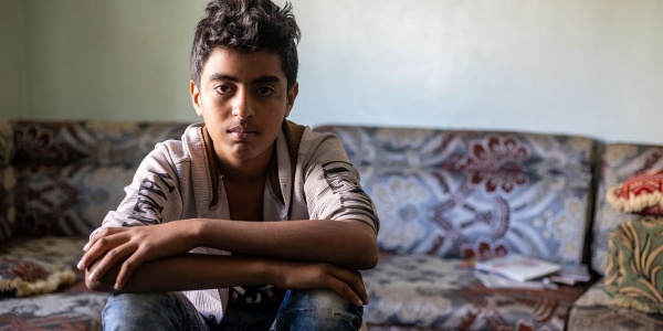 bambino yemenita seduto tiene le braccia appoggiate sulle gambe incrociate. Indossa maglietta beige e jeans