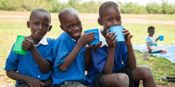 tre bambini ugandesi vestiti di blu, seduti su un ramo bevono acqua da alcune tazze colorate