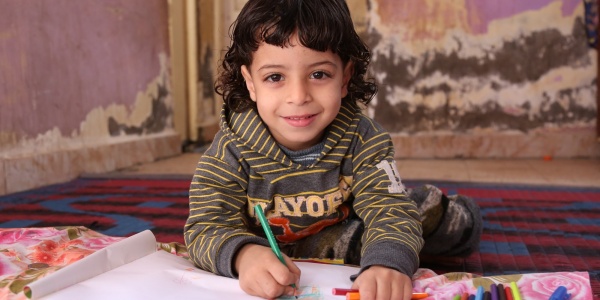 bimba egiziana con capelli neri a caschetto alla scrivania mentre disegna su un foglio bianco e guarda sorridente in camera