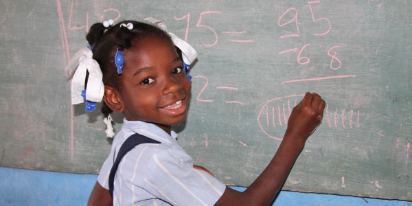 bimba che scrive alla lavagna a scuola e ci guarda con un grande sorriso. Vestita con una camicia azzurra ha un nastro tra i capelli pettinati con due treccine.